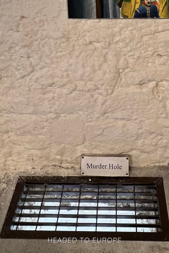 A grated metal opening labeled “Murder Hole” set into a stone floor inside an Irish castle, historically used as a defensive feature above doorways.