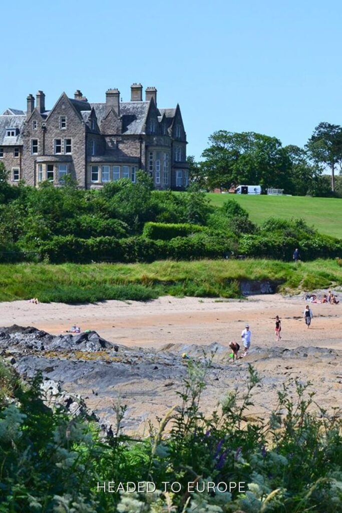 A grand stone mansion sits on a lush green hill above a sandy beach with a few people walking and playing. The sky is clear and bright blue, capturing a sunny summer day.