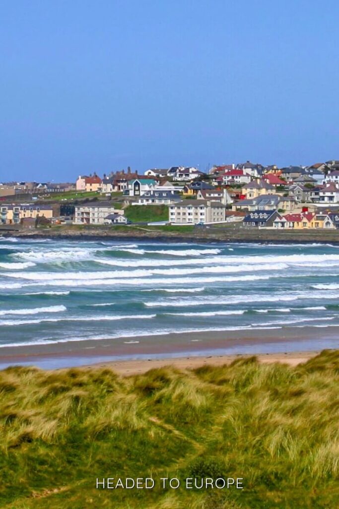 A wide sandy beach with gentle waves lapping the shore, looking out across the water to a town filled with tightly packed houses under a bright blue sky.