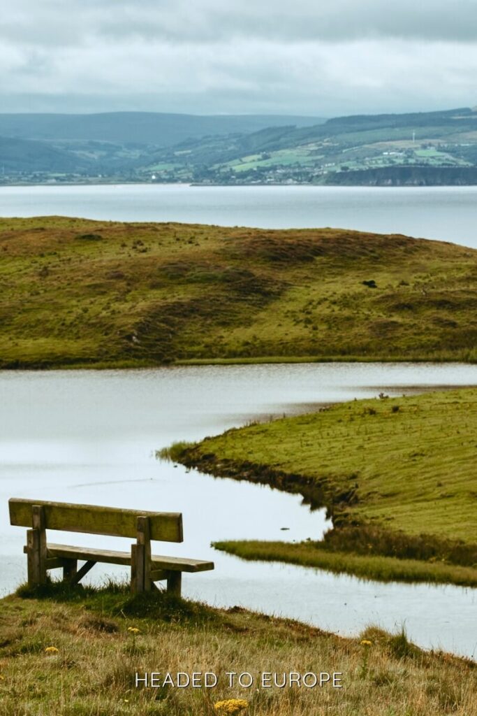 A simple wooden bench faces a peaceful lake surrounded by grassy hills, with distant views of a village and forested slopes across the water. The overcast sky adds a calm, reflective mood.