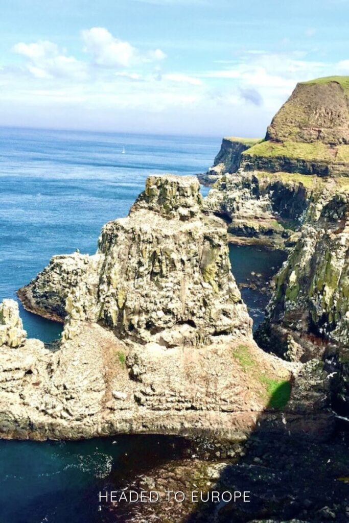 Jagged sea cliffs and rocky outcrops rise dramatically from deep blue waters along the rugged coastline of Rathlin Island, topped with bright green grass and under a clear sky.