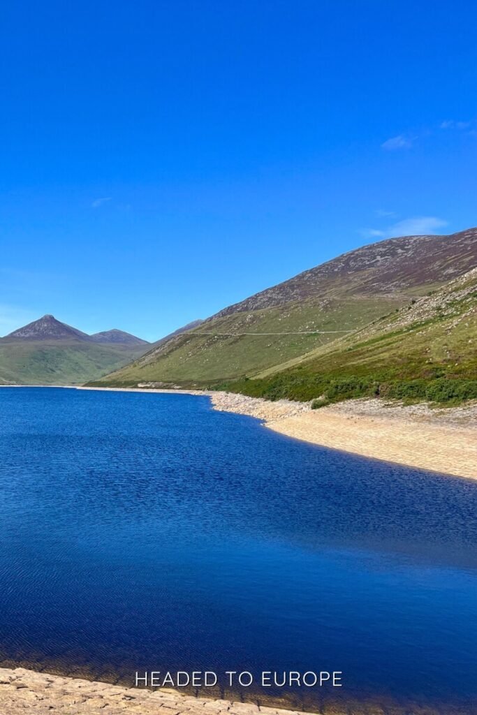 A tranquil reservoir with strikingly blue water curves along the base of green and brown hills. Under a cloudless sky, the peaks rise in the distance creating a classic Mourne Mountains landscape.