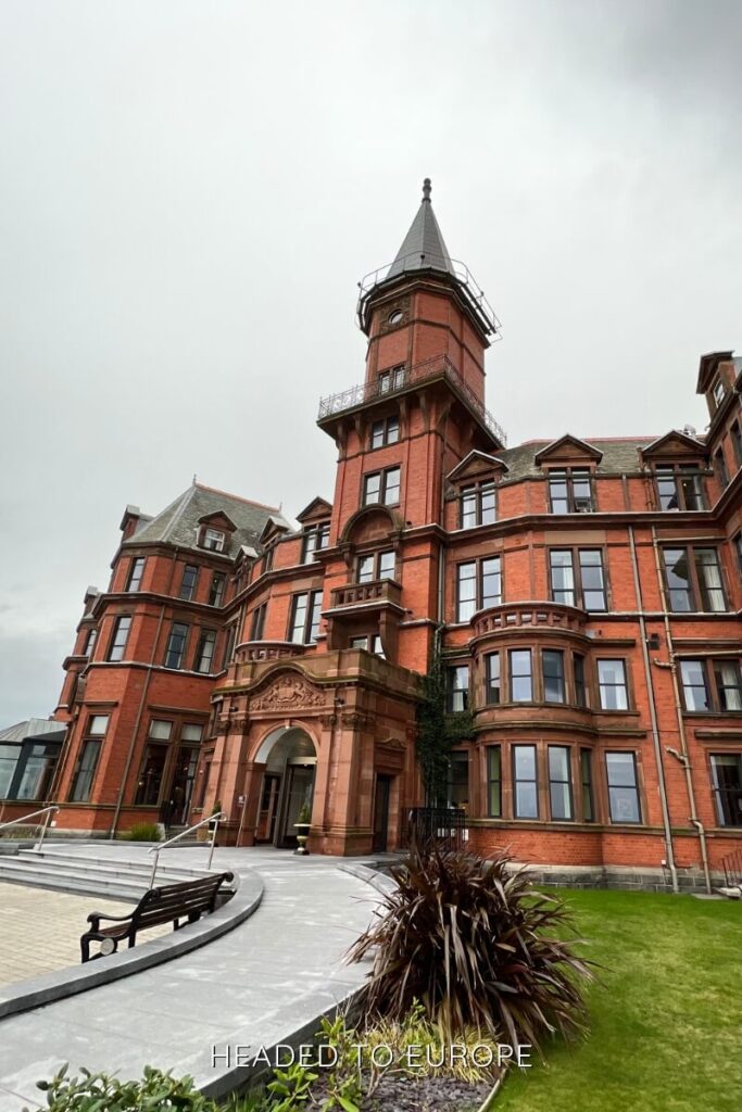 An elegant redbrick building with ornate towers and arched windows, likely a hotel, stands against a grey sky. The landscaping includes trimmed grass, a curved walkway, and benches.
