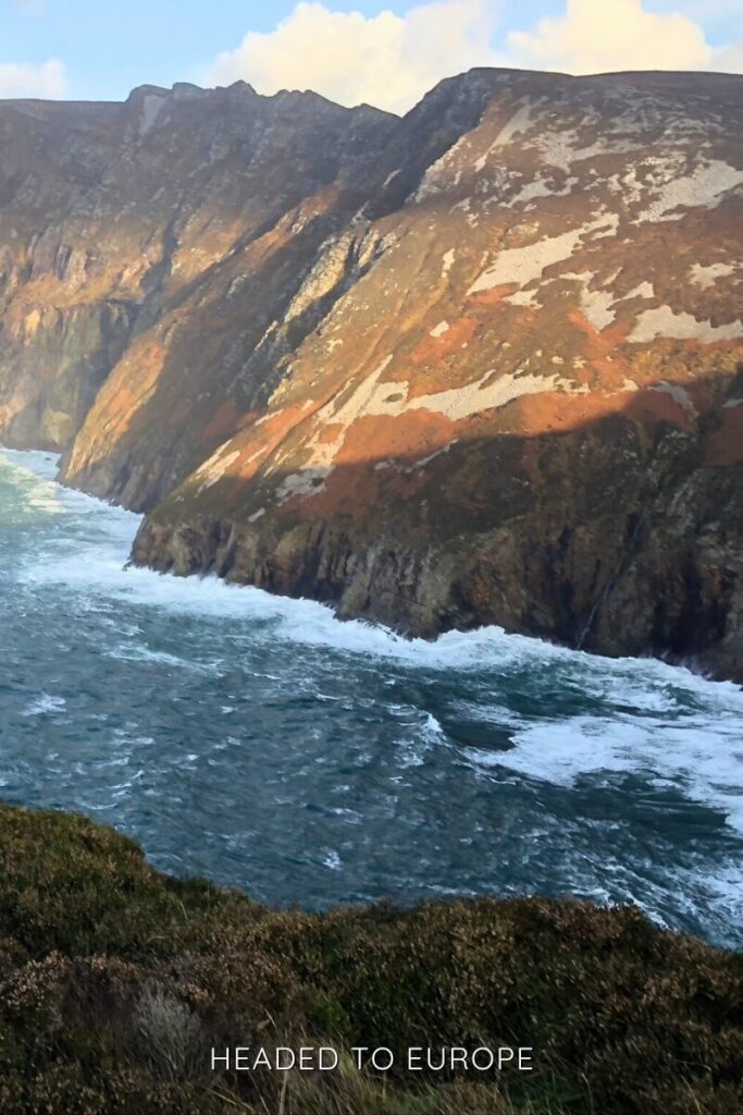 Rugged cliffs along the Atlantic coastline catch the soft light of sunset. The waves crash below, and the slopes are streaked with rust, white, and green tones.