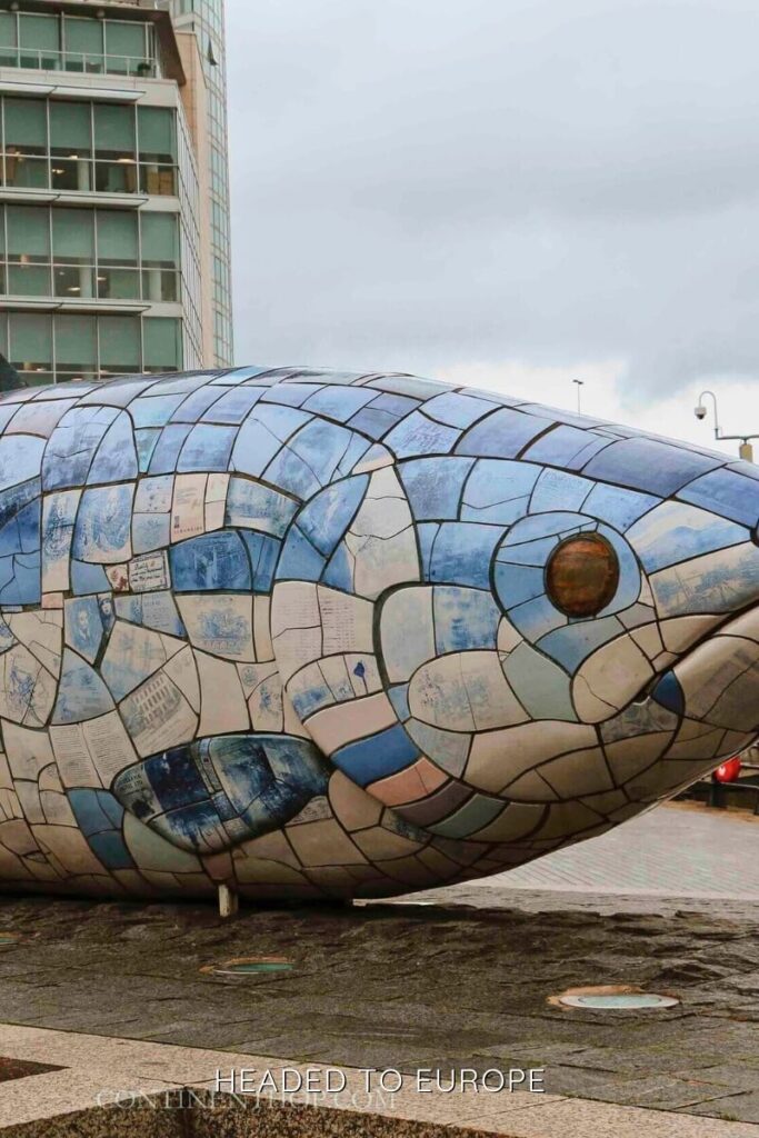 A close-up of the Salmon of Knowledge sculpture in Belfast, made of blue, white, and ceramic tiles that resemble fish scales. Modern buildings rise in the background under a cloudy sky.