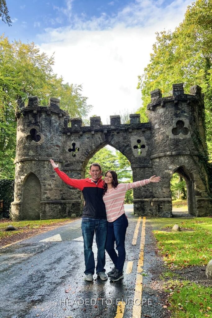 A smiling couple with arms outstretched stands on a tree-lined road in front of a fairytale-like stone gatehouse with arched doorways and castle-style turrets.