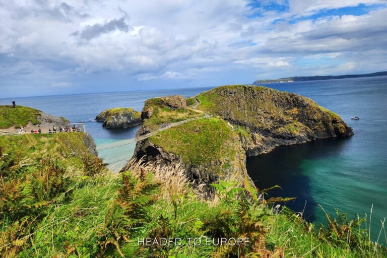 A scenic coastal view of the Carrick-a-Rede Rope Bridge stretching between two grassy cliffs over turquoise waters, with visitors gathered on one side and a walking trail winding up the rocky island ahead under a partly cloudy sky.